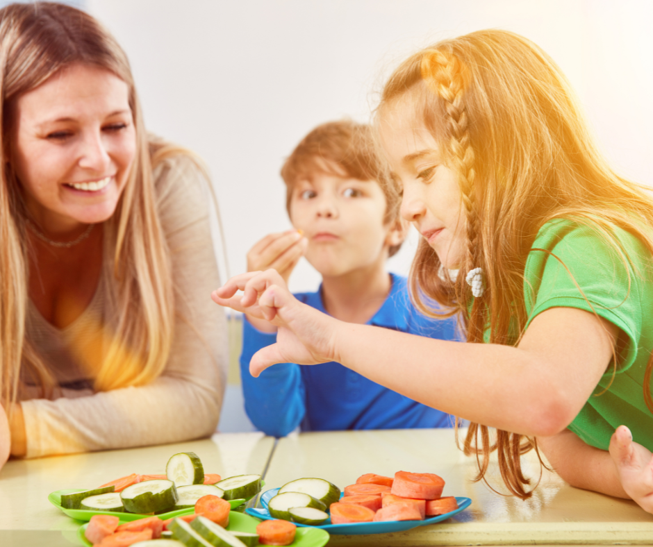 Niña eligiendo rodajas de vegetales frescos mientras un niño prueba uno y una mujer adulta sonríe, en un ambiente de alimentación saludable y aprendizaje familiar.