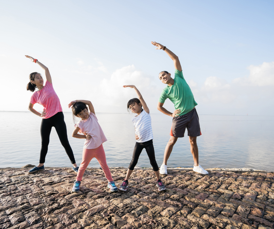 Familia haciendo ejercicio al aire libre junto al mar, con padres e hijas estirándose de forma sincronizada, promoviendo la actividad física y el bienestar en familia.