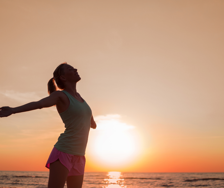 Mujer disfrutando del aire libre al atardecer junto al mar, con los brazos abiertos en señal de libertad y bienestar.