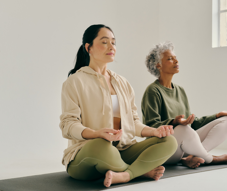 Dos mujeres de distintas edades practicando meditación sentadas en postura de loto, con ojos cerrados y expresión de calma.