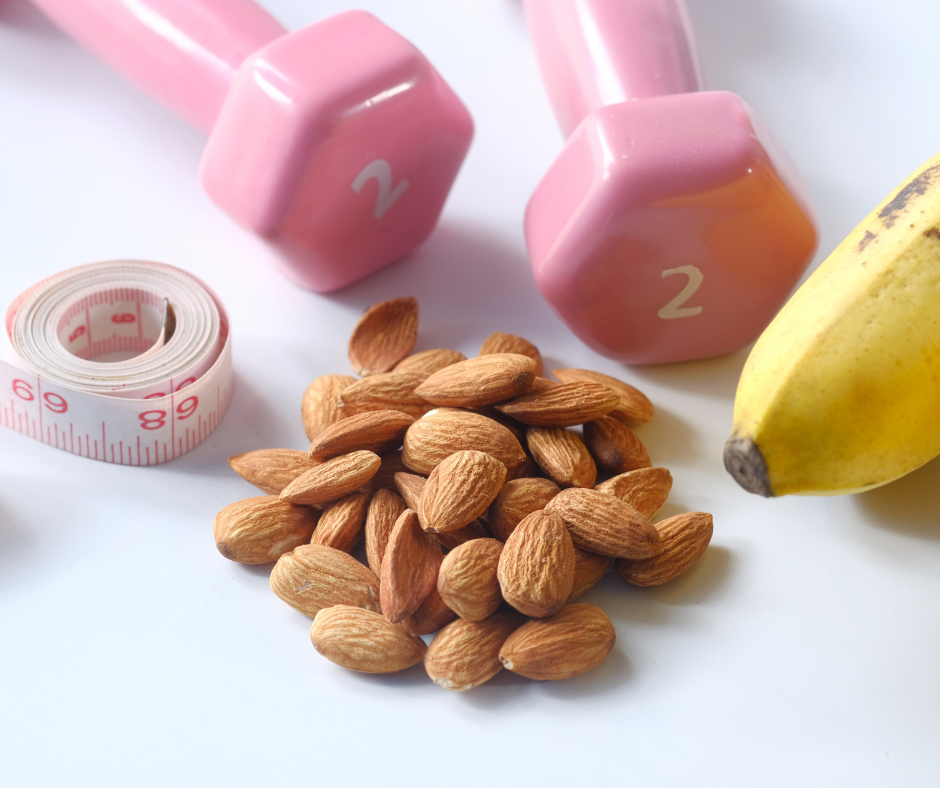 Almendras, una banana, cinta métrica y dos mancuernas rosas sobre una superficie blanca, representando hábitos de alimentación saludable y ejercicio.