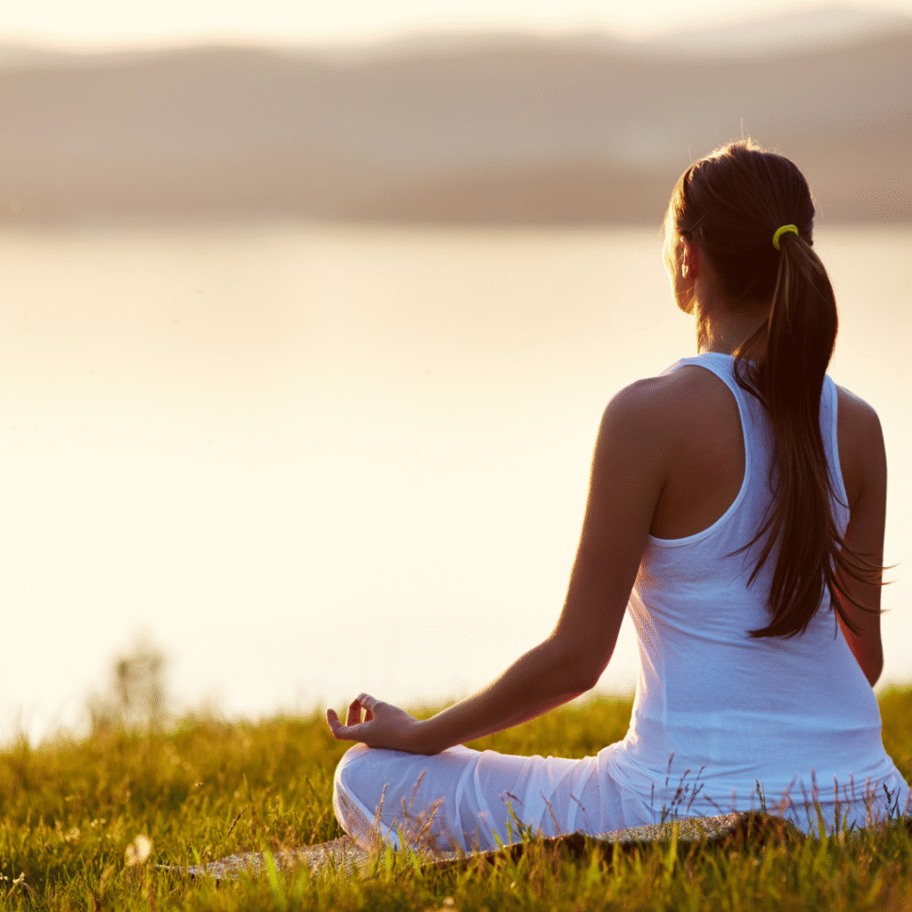 Mujer joven practicando meditación al aire libre, sentada en postura de loto sobre el pasto al atardecer, con ropa blanca, representando calma, bienestar y mindfulness
